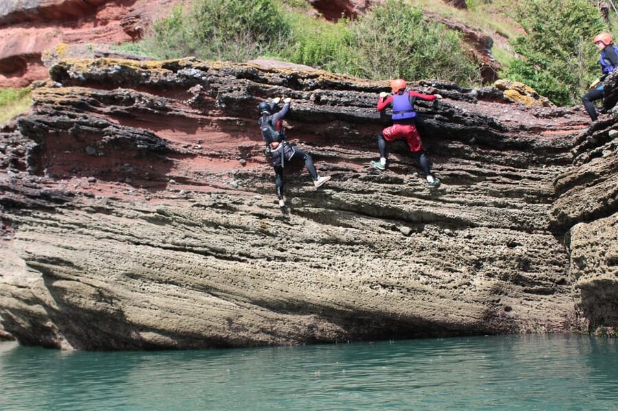 Torquay: Coasteering 5<em> Geopark Tour in Torquay – Meeting the orange van at The Thatched Tavern’ /></p>
<p><p>The meeting point is outside The Thatched Tavern pub in Maidencombe. Look for the orange van. Parking is available nearby, but parking fees are not included, and you must arrange your own transport to and from the meeting point.</p>
</p>
<p><p>This meeting point matters because the tour is outside central Torquay. Give yourself time to reach Maidencombe, find the pub, and arrive ready for the briefing. Do not count on turning up in ordinary holiday clothes and starting at once. You need time to change and get fitted with equipment.</p>
</p>
<p><p>Once you arrive, the team provides the main safety kit:</p>
</p>
<ul>
<li>Wetsuit</li>
<li>Helmet</li>
<li>Buoyancy aid</li>
<li>Gloves</li>
<li>Old shorts</li>
<li>Secure storage for belongings</li>
</ul>
<p><p>Bring your own swimwear, towel, change of clothes, sunscreen, and long socks. You also need old trainers or water shoes. Water shoes are suitable, and old trainers can be borrowed if needed, but it is still wise to ask about footwear when booking. Your shoes will get wet and may scrape against rock, so do not bring your best pair.</p>
</p>
<p><p>A wetsuit helps with the cold, but it does not turn the sea into a warm swimming pool. If you are sensitive to cold water, the swimming sections may feel demanding even with the provided gear.</p>
</p>
<h2>The safety briefing comes first</h2>
<p><img width="900" height="599" decoding="async" src=